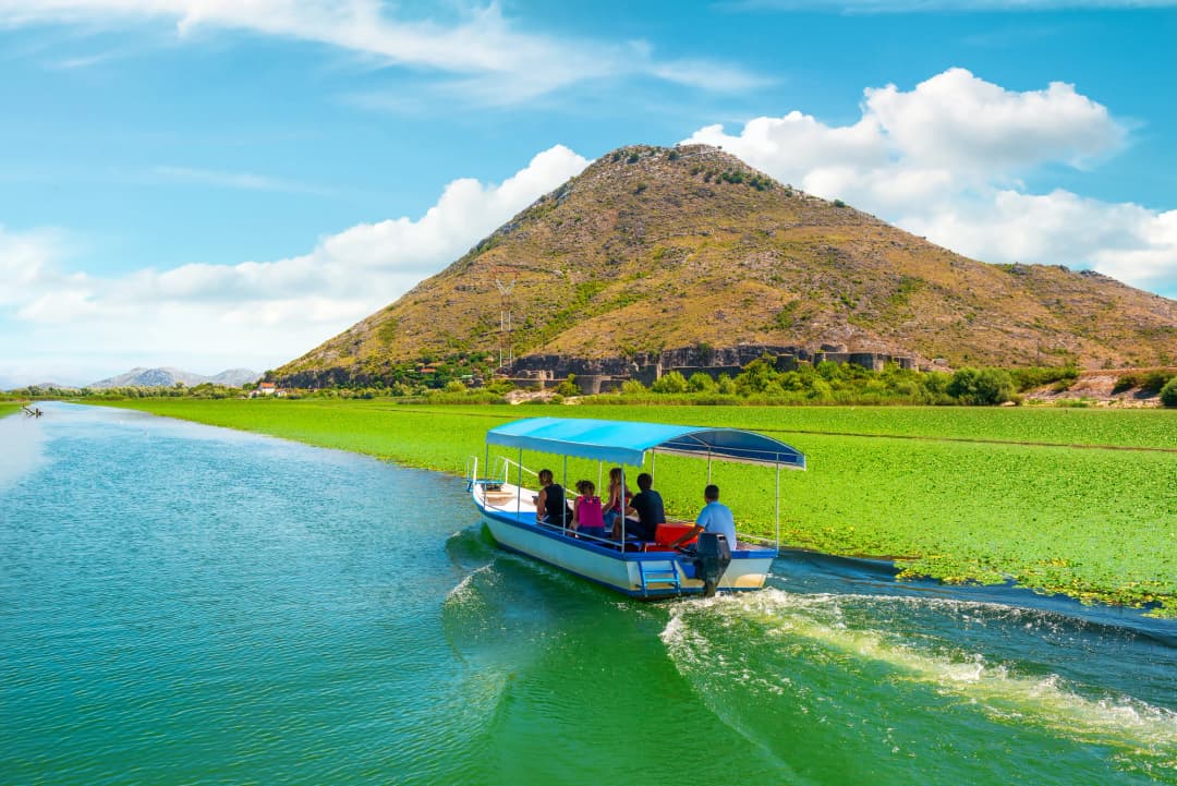 A small visitor boat passing water lilies below a wooded Lake Skadar hillside