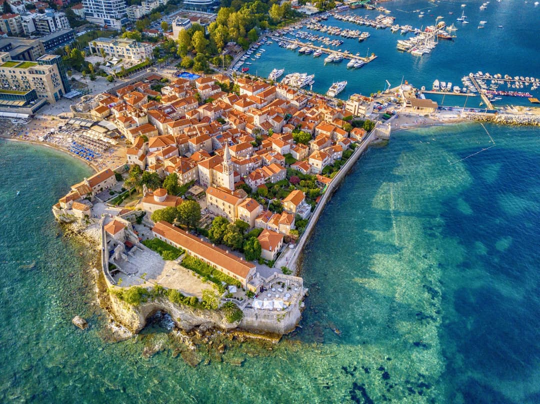 Pogled iz vazduha na poluostrvo budvanskog Starog grada koje zalazi u tirkizni Jadran, sa zbijenim naseljem u bedemima i plažama uokolo