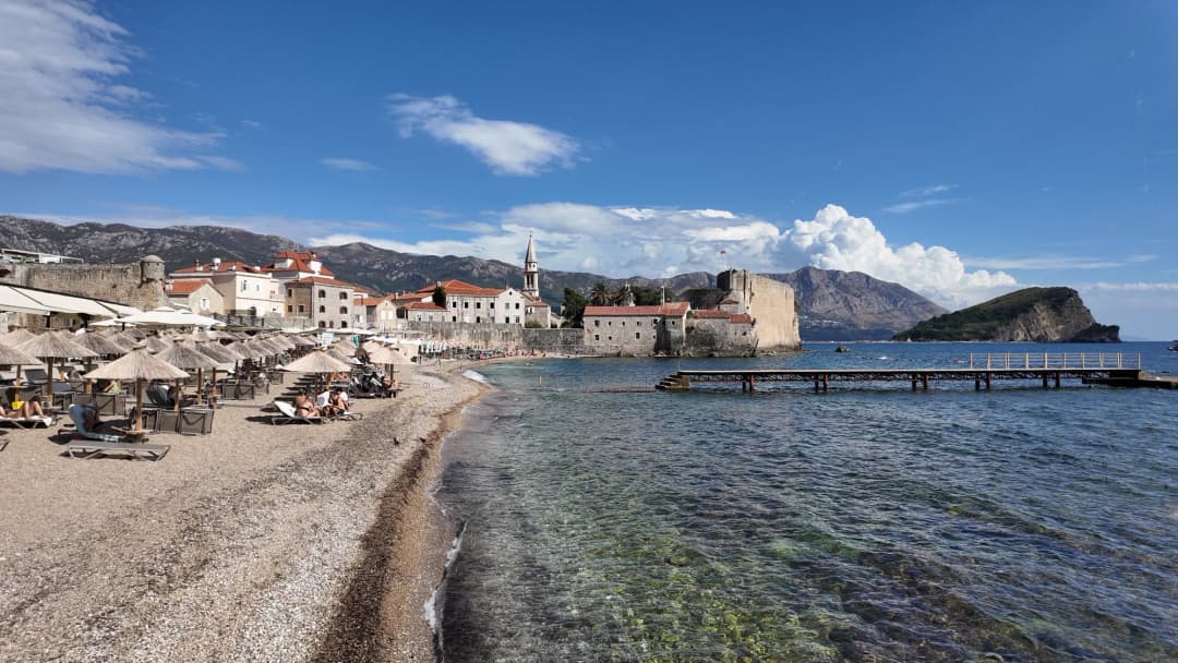 Plaža Ričardova glava puna ležaljki i suncobrana od trske, tik ispod zidina Starog grada, a u pozadini ostrvo Sveti Nikola.