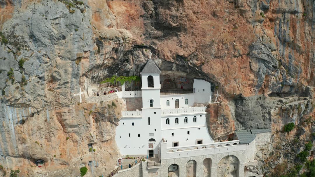 Okrečena fasada manastira Ostrog uklopljena u strmu sivu liticu visoko iznad Bjelopavlićke doline.