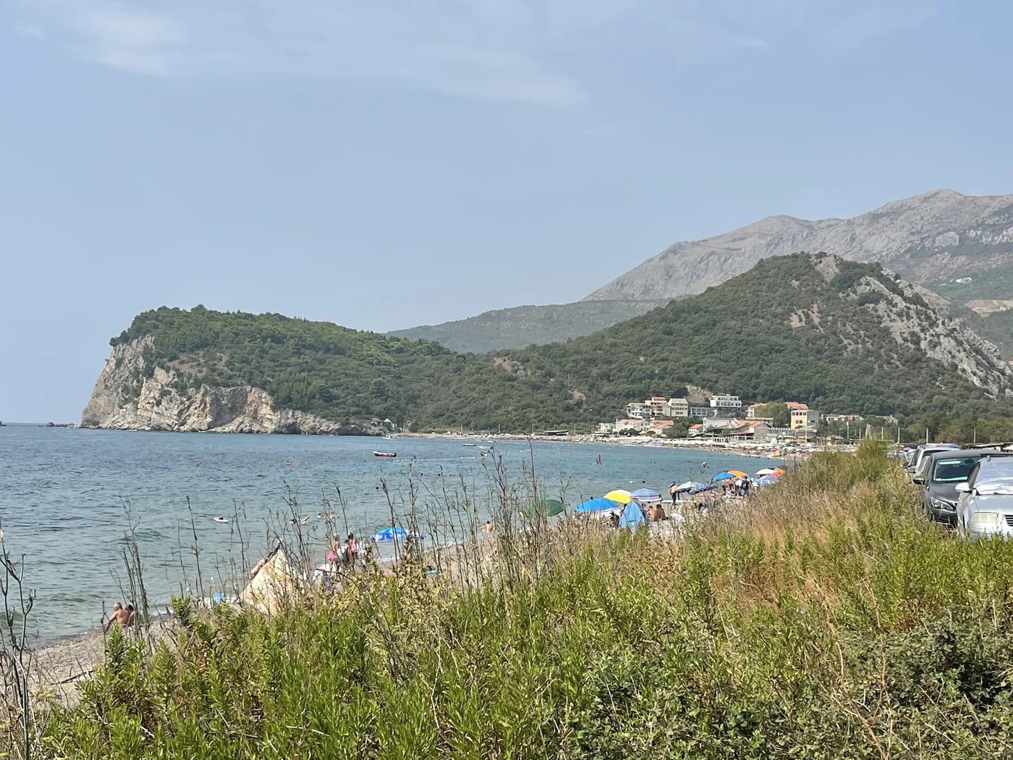 Buljarica from the track behind the sand: umbrellas along a 2 km arc of grey sand and shingle, campervans parked in the scrub of the wetland, the headland and the Paštrovići hills beyond