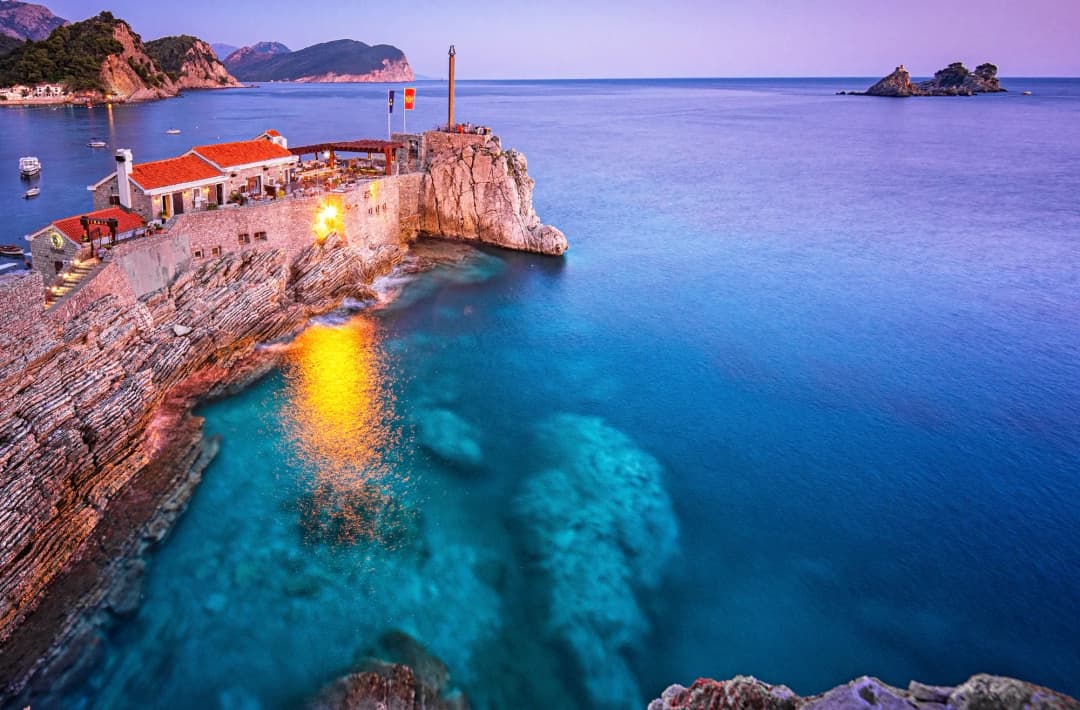 Tvrđava Castello na svojoj stijeni u Petrovcu u sumrak, sa svjetlima koja se ogledaju u tirkiznoj vodi i ostrvcima na horizontu