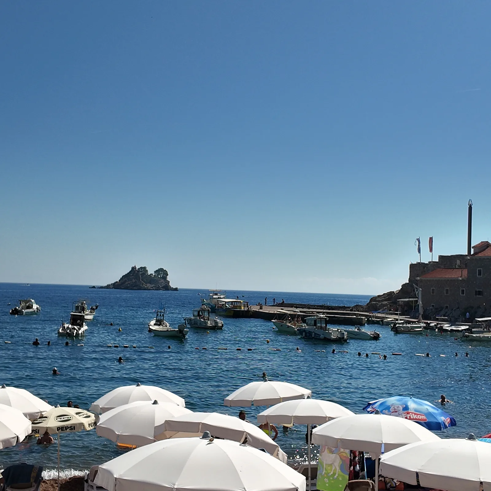Bijeli suncobrani na sjevernom kraju petrovačke plaže, kupači unutar kupališta obilježenog bovama, mali čamci privezani kod kamenog gata, tvrđava Castello na svojoj stijeni desno i ostrvca na horizontu lijevo