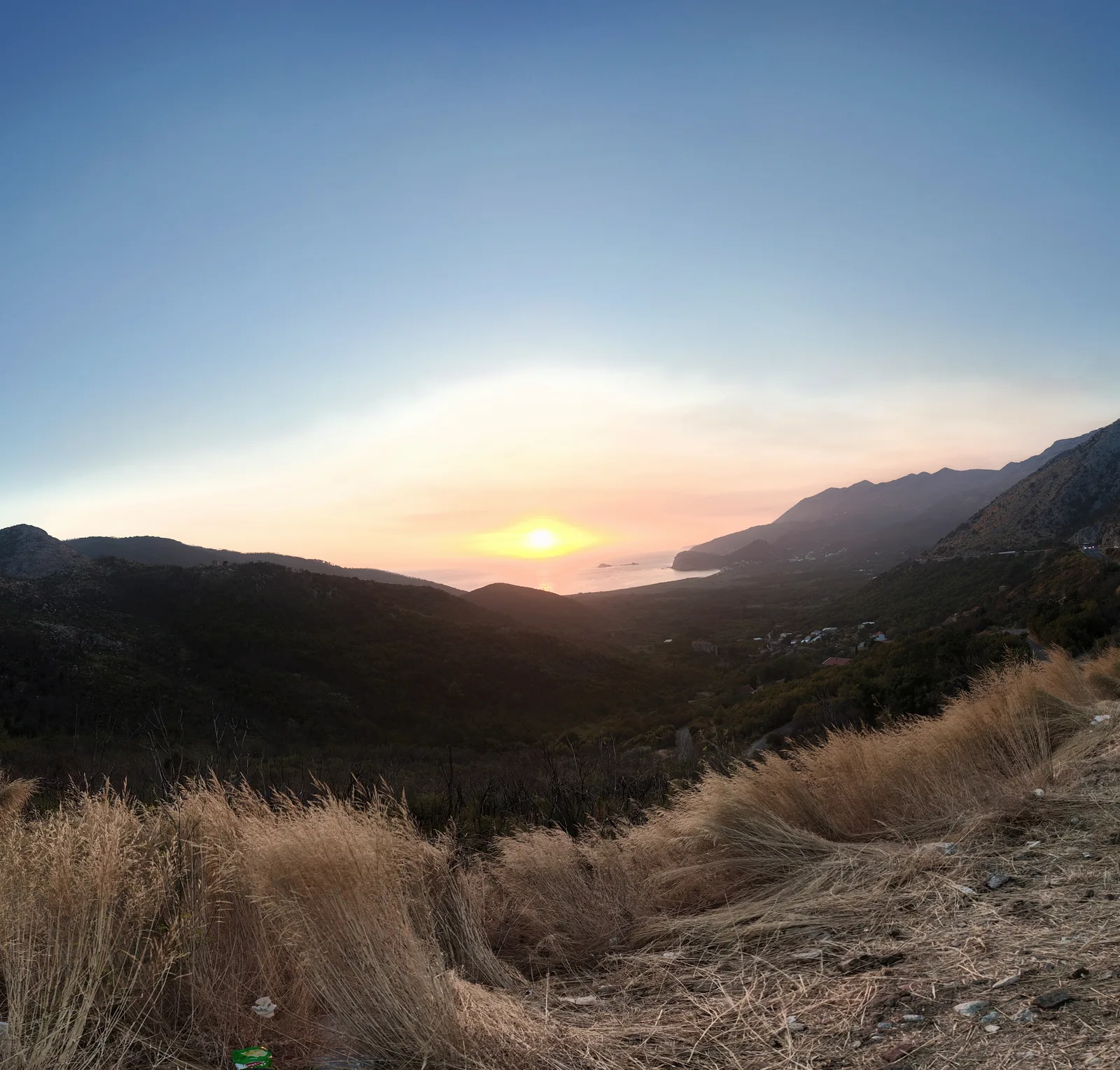 Zalazak sunca nad Jadranom s obalnog puta iznad Buljarice — sunce nisko nad morem, dugi luk buljaričke plaže i njena zelena ravnica ispod, a ostrvca Katič vidljiva iza rta prema Petrovcu