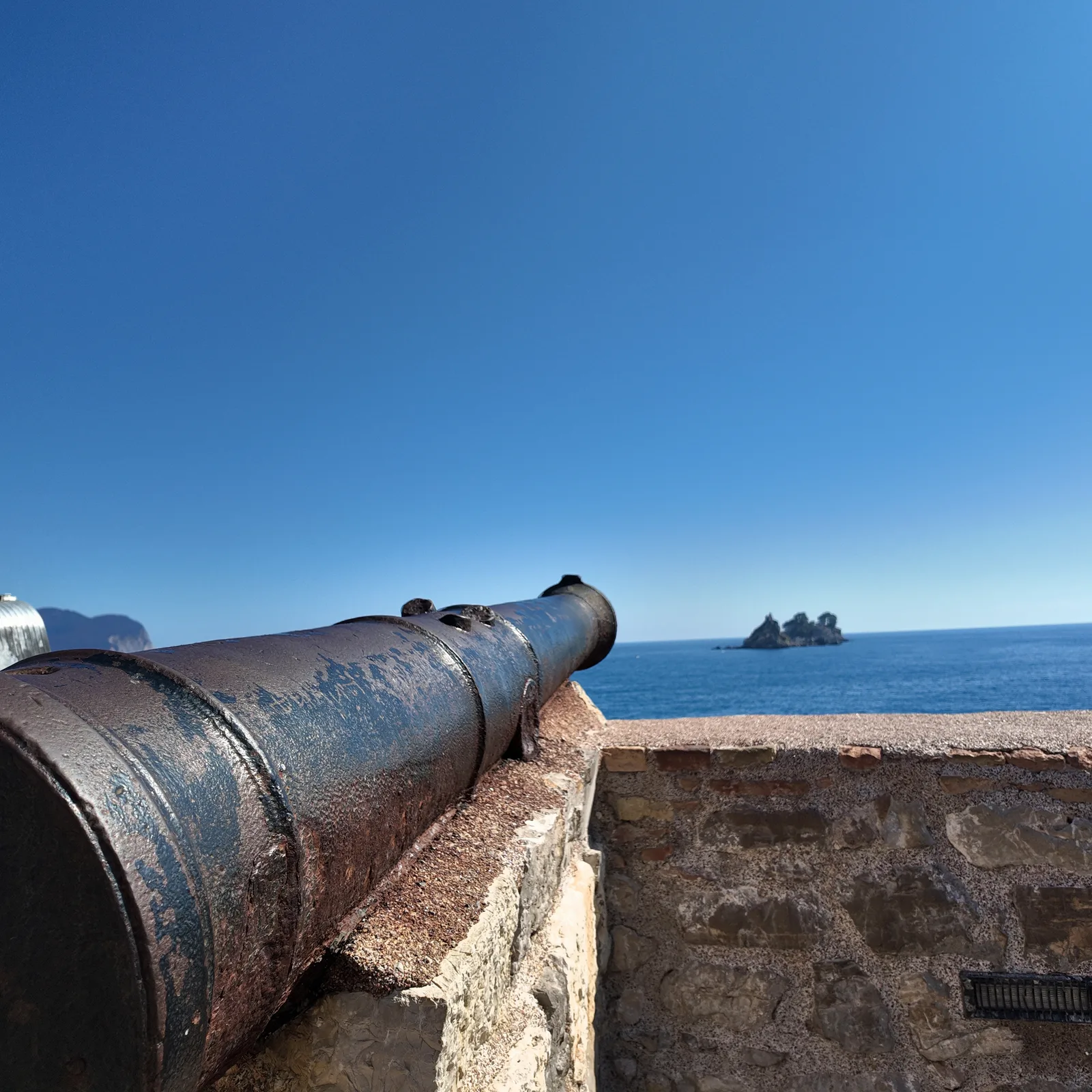 Zarđali gvozdeni top na kamenom parapetu tvrđave Castello u Petrovcu, uperen prema tamnoplavom moru, sa pošumljenim ostrvcima u daljini desno